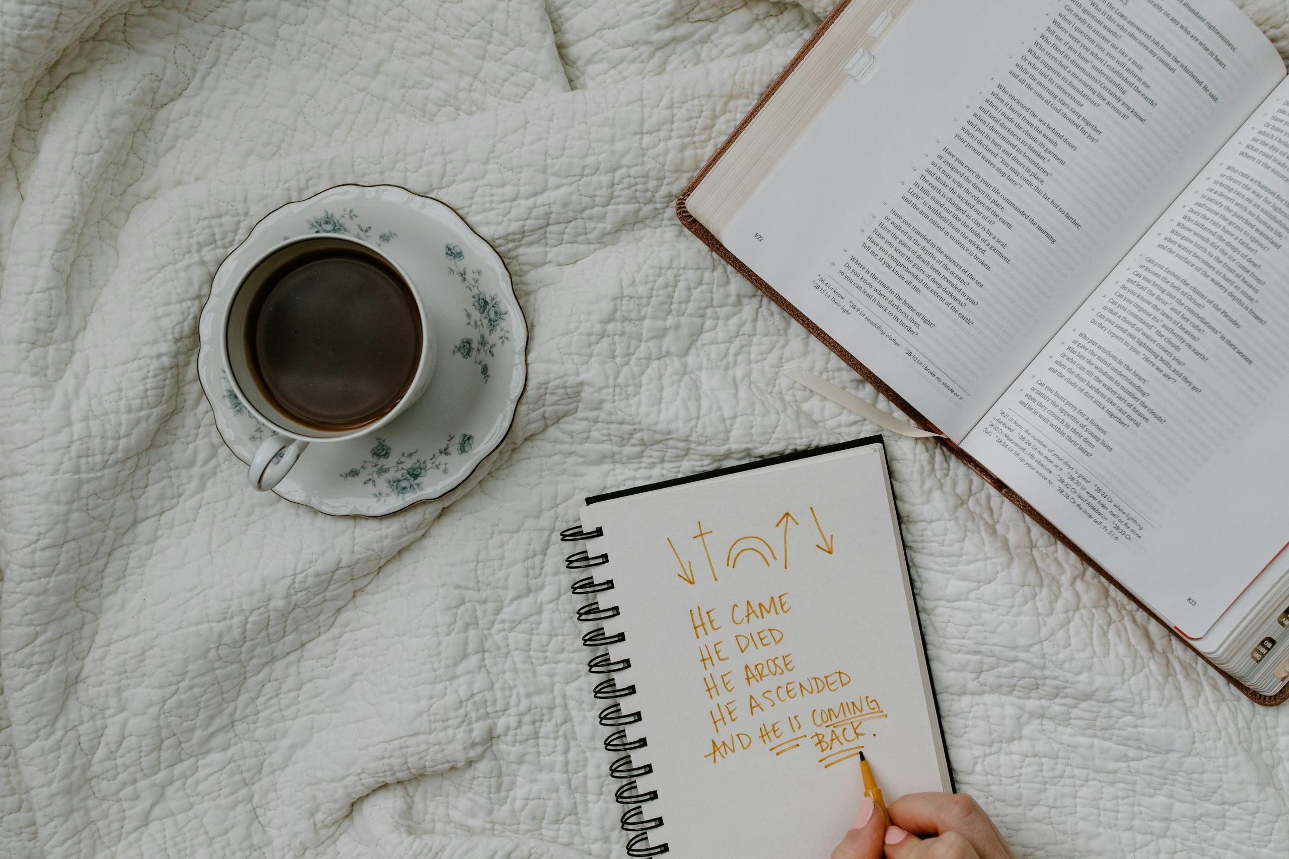 A Person Writing on the Notebook Beside a Book and Coffee